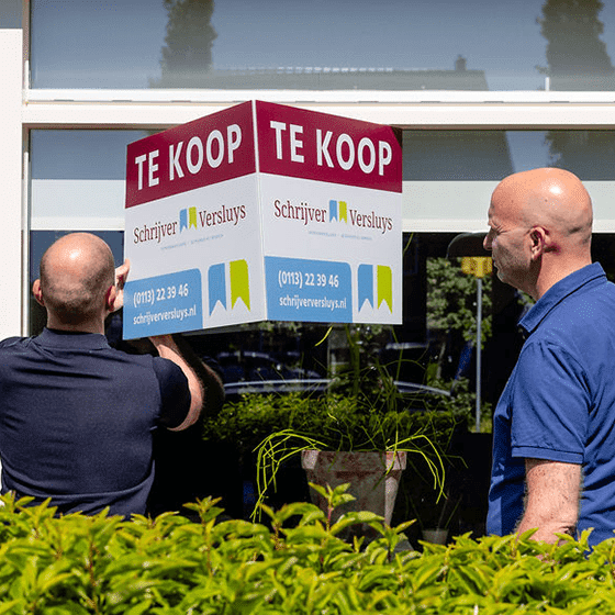 Aard-Jan en Mike, makelaars van Schrijver Versluys, hangen samen een verkoopbord op.